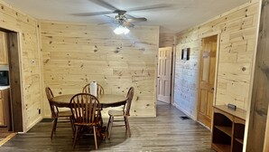A wooden interior with a dining table and chairs, a ceiling fan, and a small shelf with a sign.