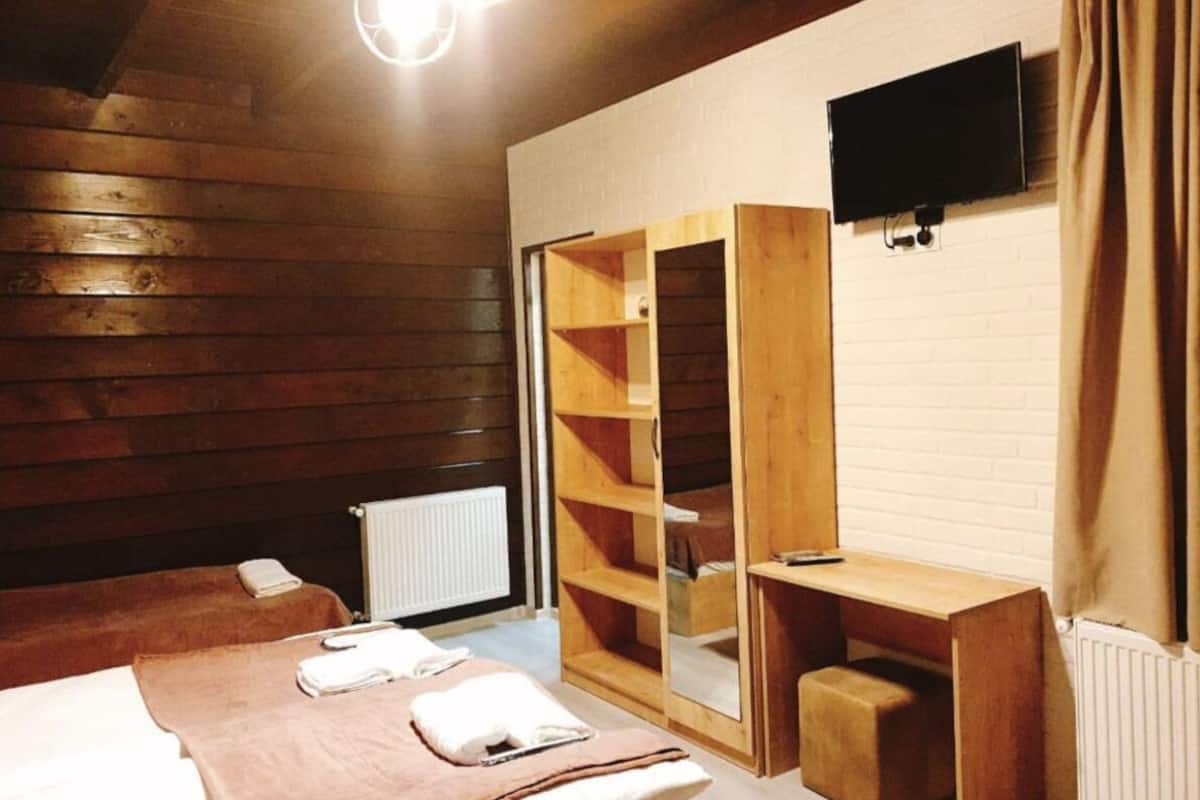 A wooden room with a bed, a wooden wardrobe, a small table, and a TV mounted on the wall.