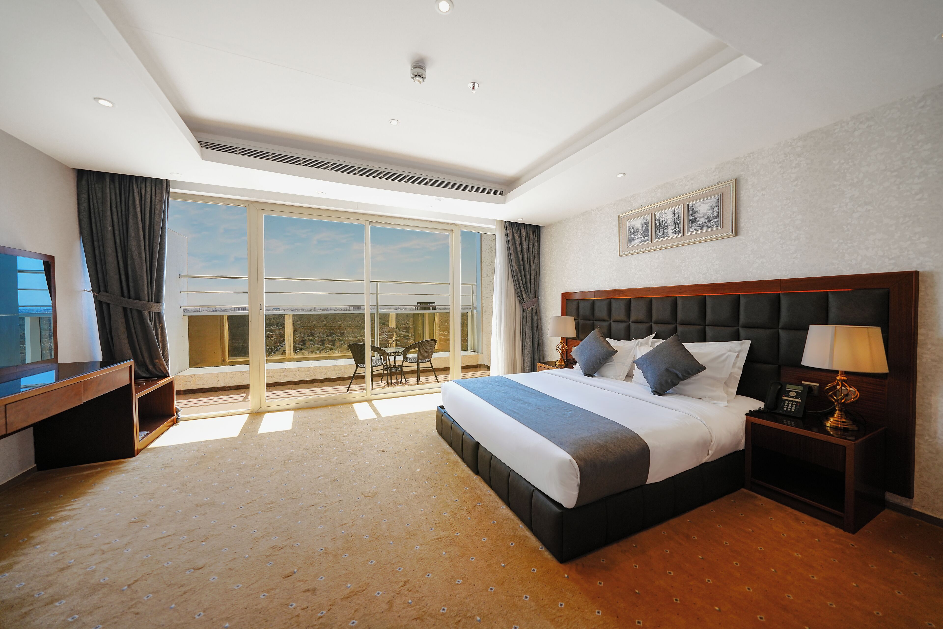 A hotel room with a large bed, a desk, a chair, a nightstand, and a view of the city through large windows.