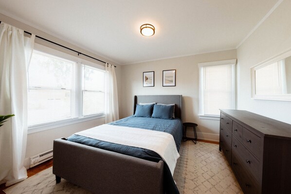 A bedroom with a bed, a dresser, and two windows with blinds.