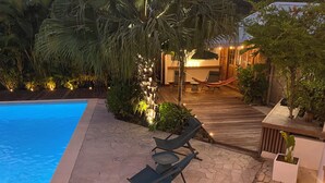 A poolside area with a stone patio, lounge chairs, a hammock, and a wooden deck leading to a building with warm lighting. - La Villa d'à Côté (Le Marin)