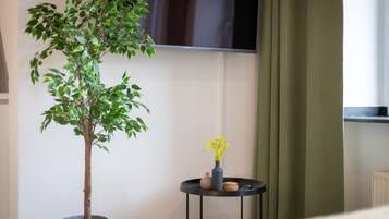 A potted tree with green leaves, a small table with a vase of yellow flowers, and a green curtain in a room with a white wall and a window.