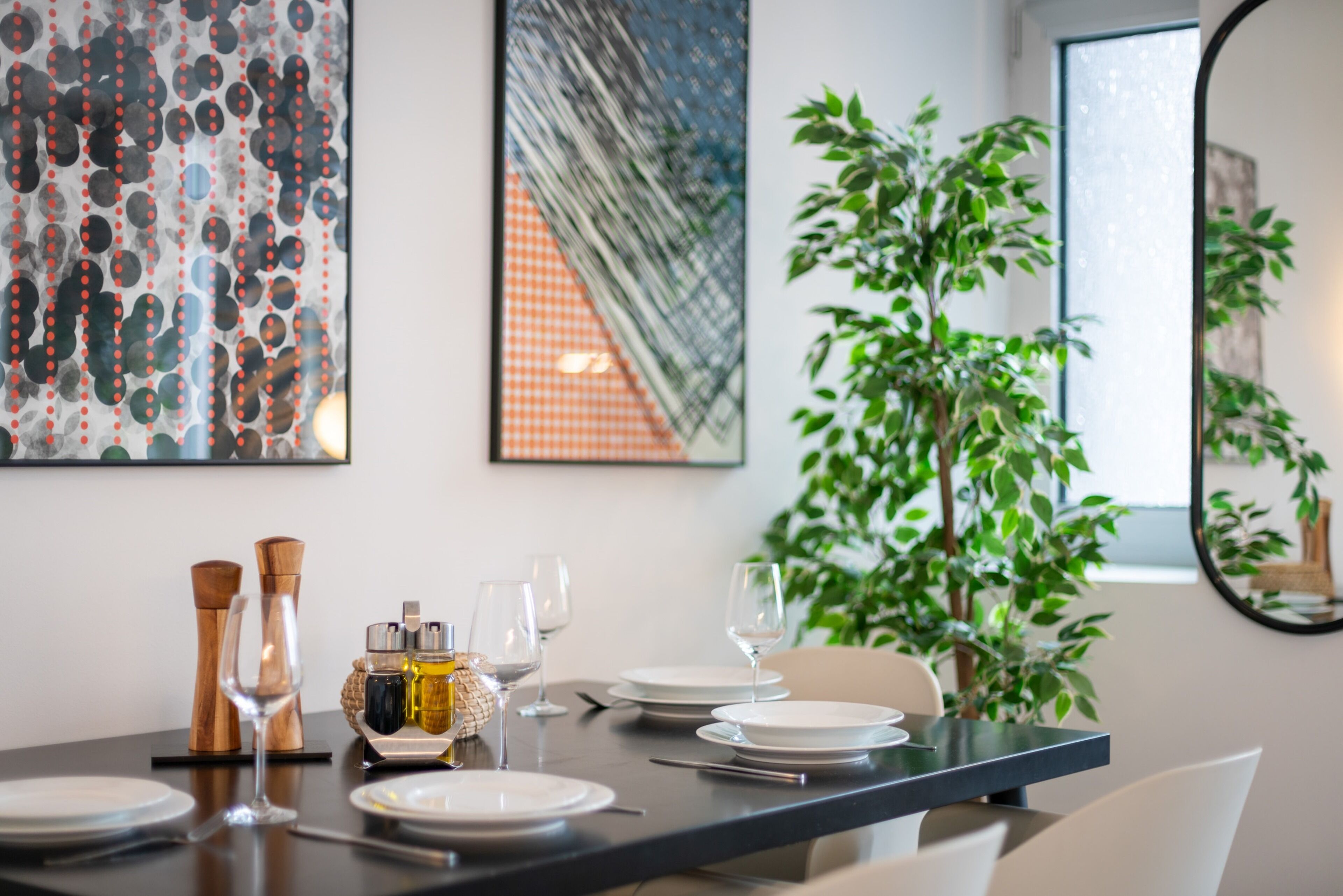 A dining area with a table set for four, featuring a modern design with a dark table surface and white plates. There are salt and pepper shakers, a bottle of olive oil, and wine glasses. A large potted plant is visible in the background, and the room has a contemporary feel with abstract art on the walls.