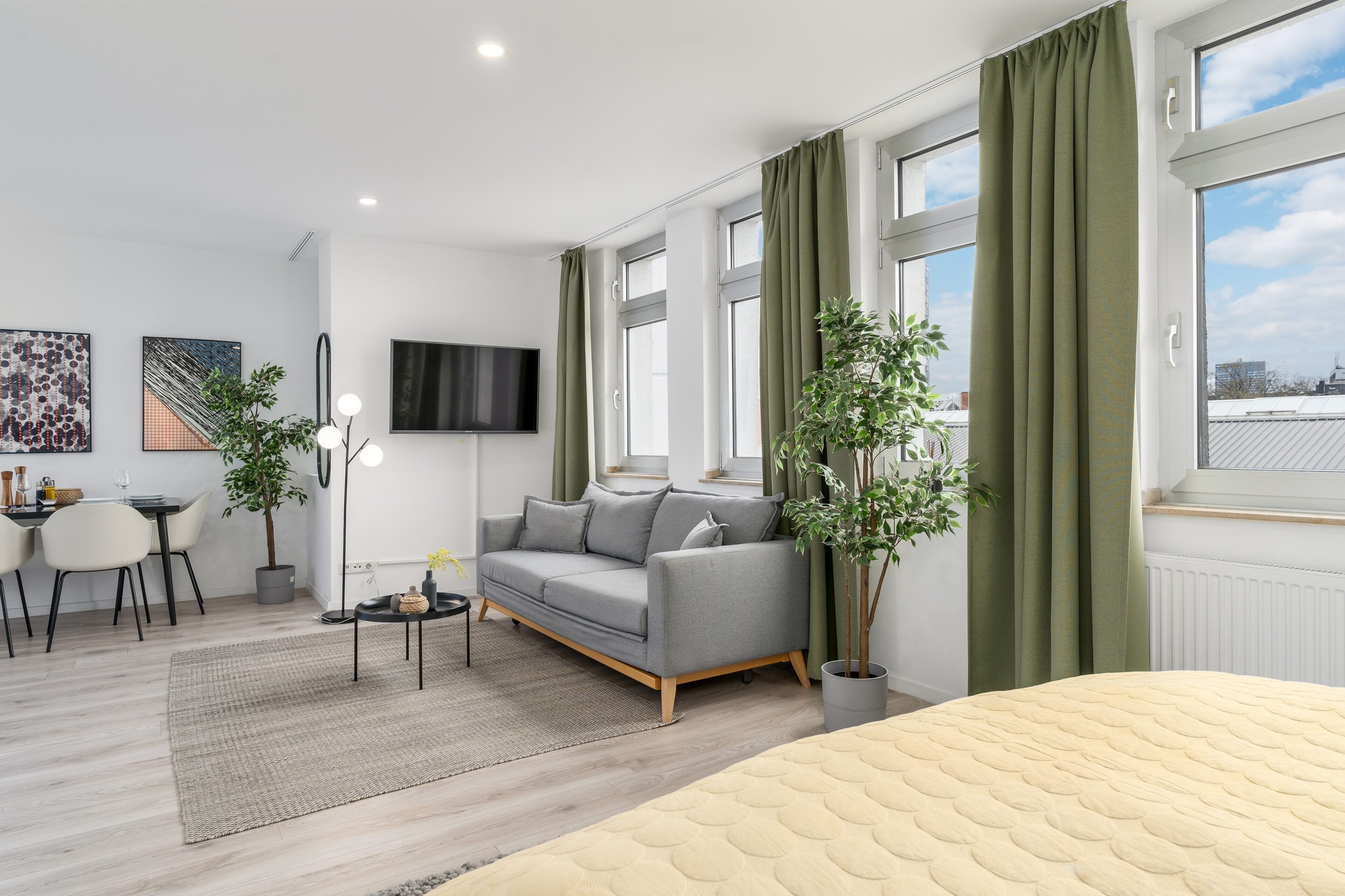 Una sala de estar moderna con un sofá gris, una cama redonda, un comedor y ventanas amplias con cortinas.