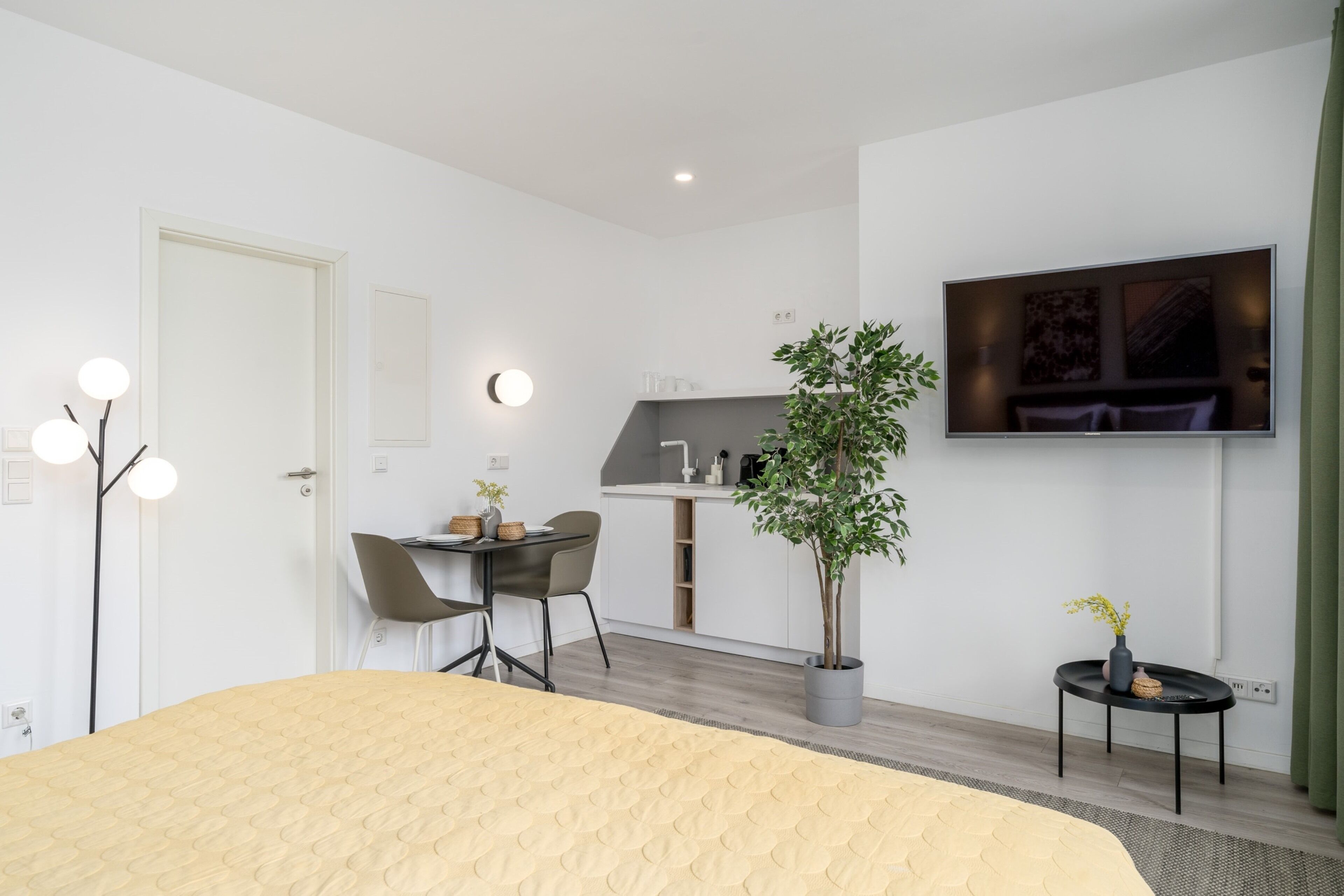 Ein modernes Hotelzimmer mit Bett, Schreibtisch, Stuhl, Zimmerpflanze, kleinem Tisch und einem an der Wand befestigten Flachbildfernseher.