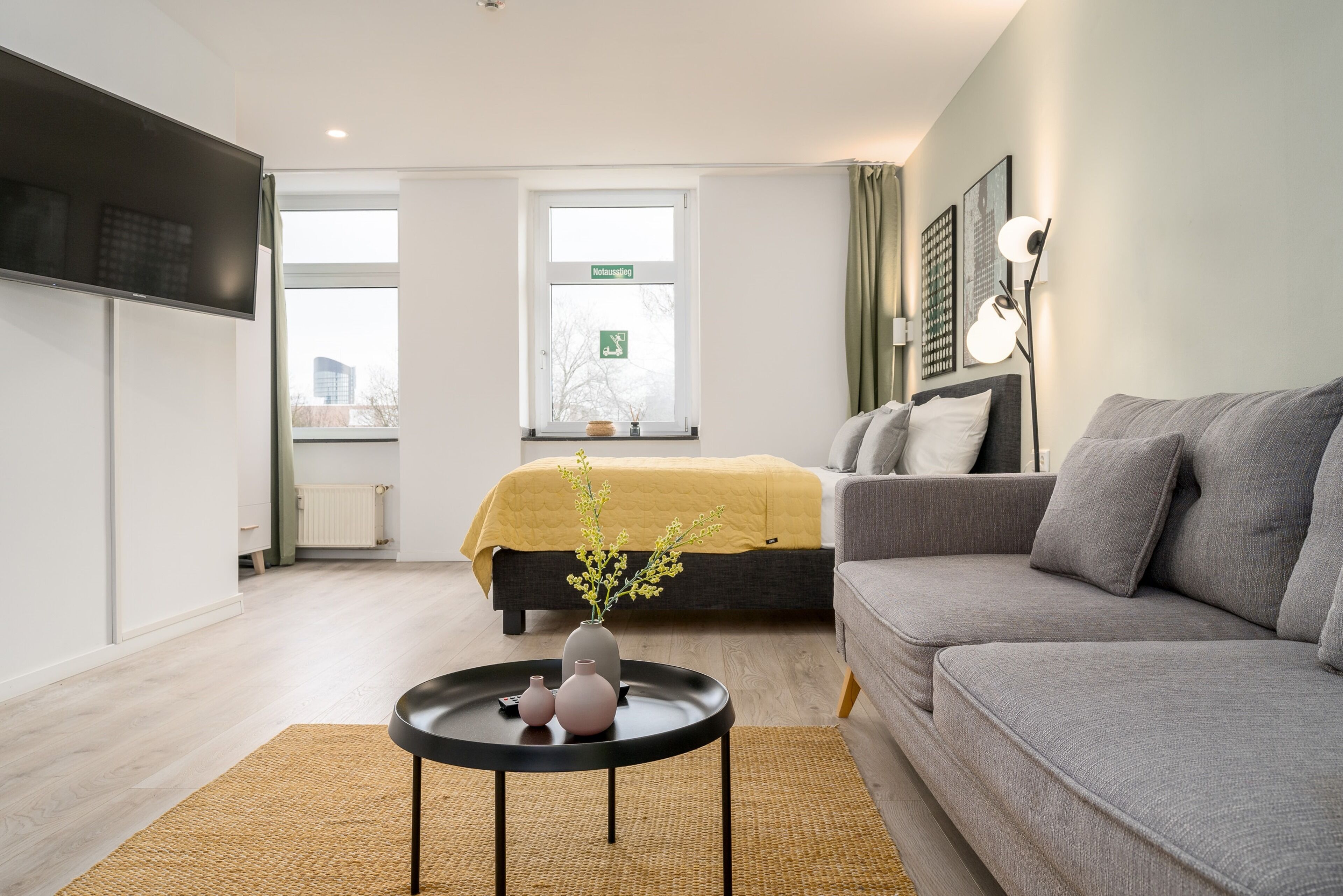 A modern hotel room with a grey sofa, a bed with a yellow blanket, a small round table with vases, and a window with a view of the city.