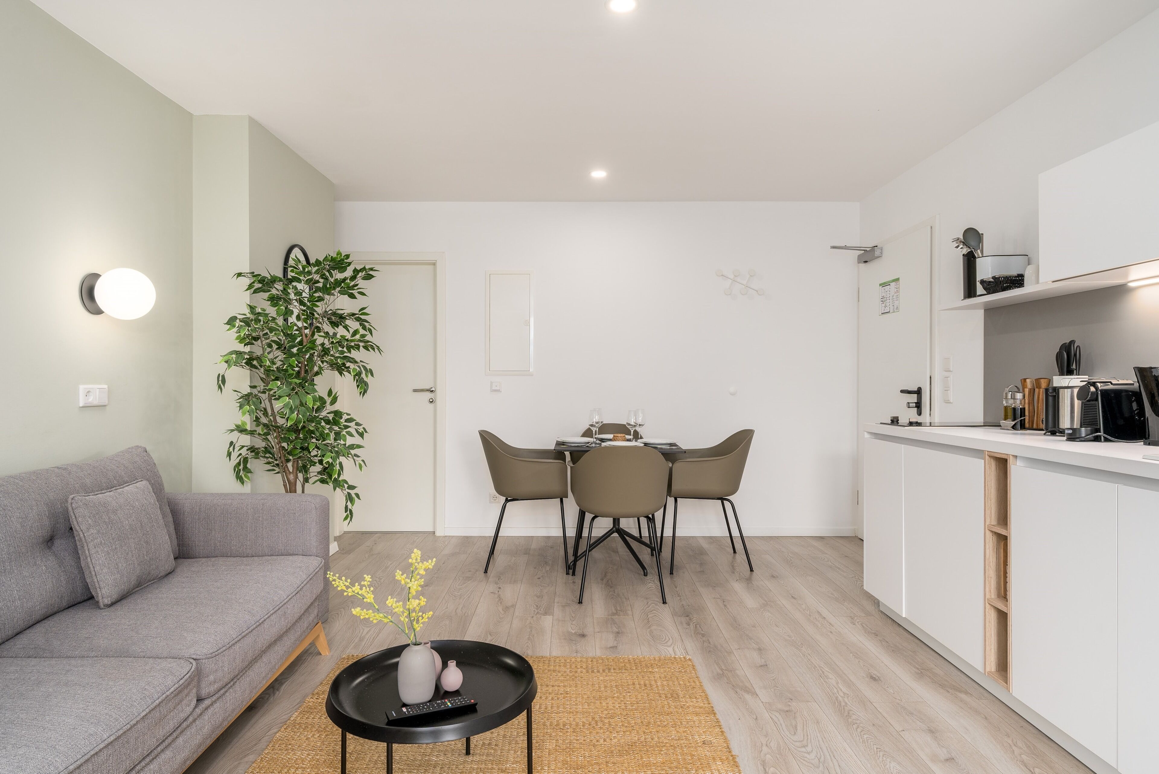 Una sala moderna con un sofá gris, una mesa de comedor con sillas, una pequeña mesa de centro y una zona de cocina con armarios blancos y una encimera.