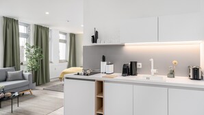 A modern kitchen with a central island, white cabinetry, and a built-in oven. There is a dining area with a grey sofa and a small table. - BRIGHT Dortmund City Center (Dortmund)