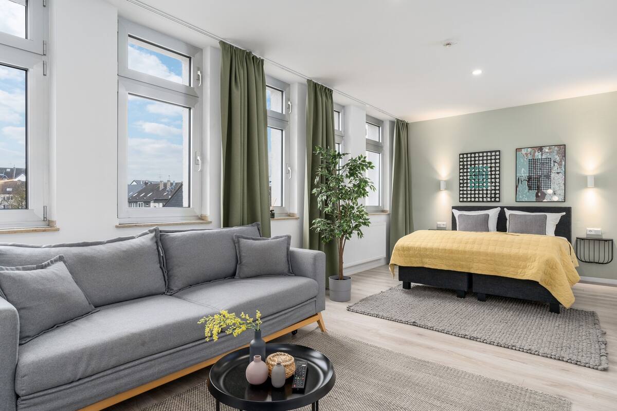 A modern living room with a grey sofa, a yellow bed, a large window, and a potted plant.