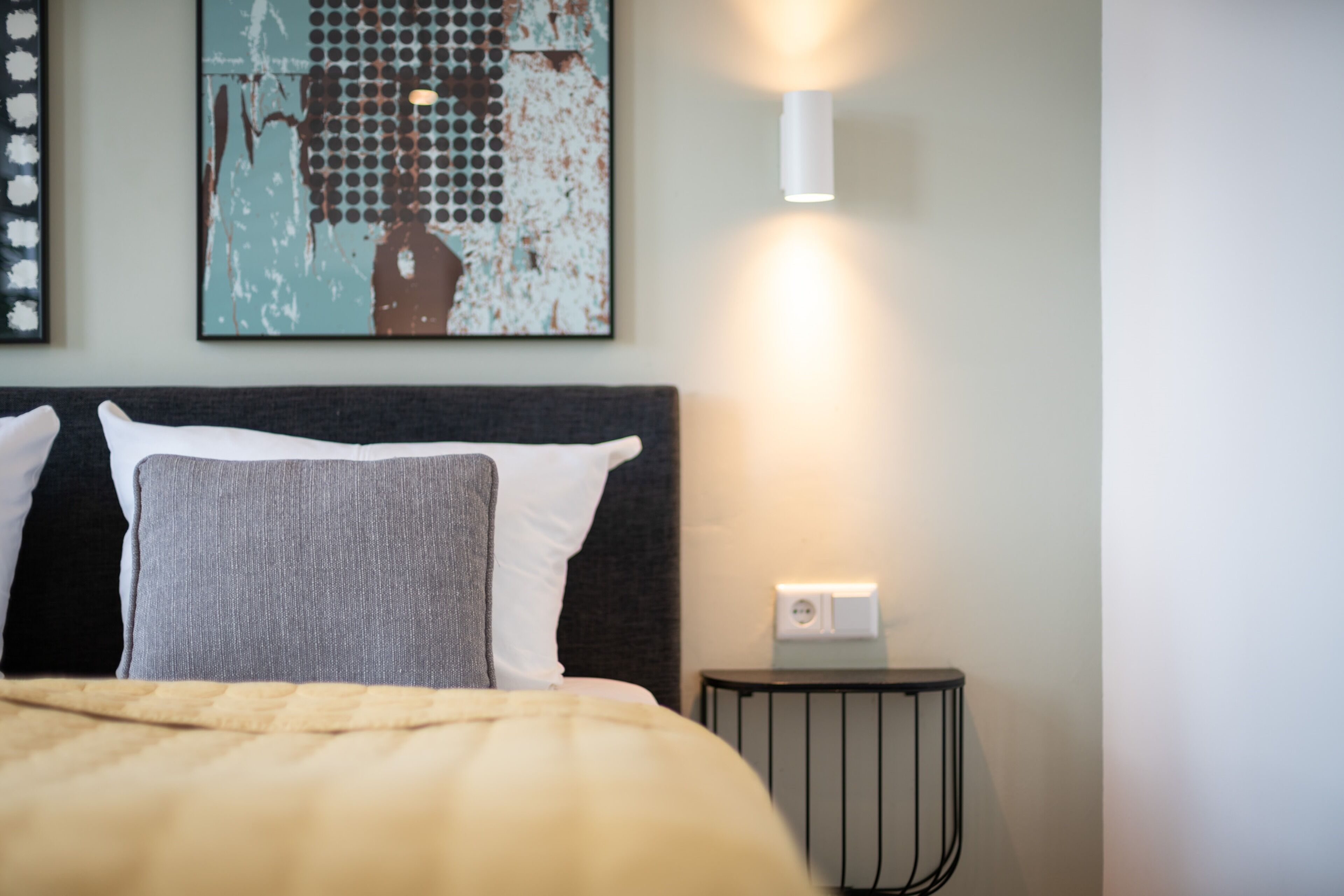 A neatly made bed with a gray and white pillow, a small bedside table with a lamp, and a wall-mounted light fixture.