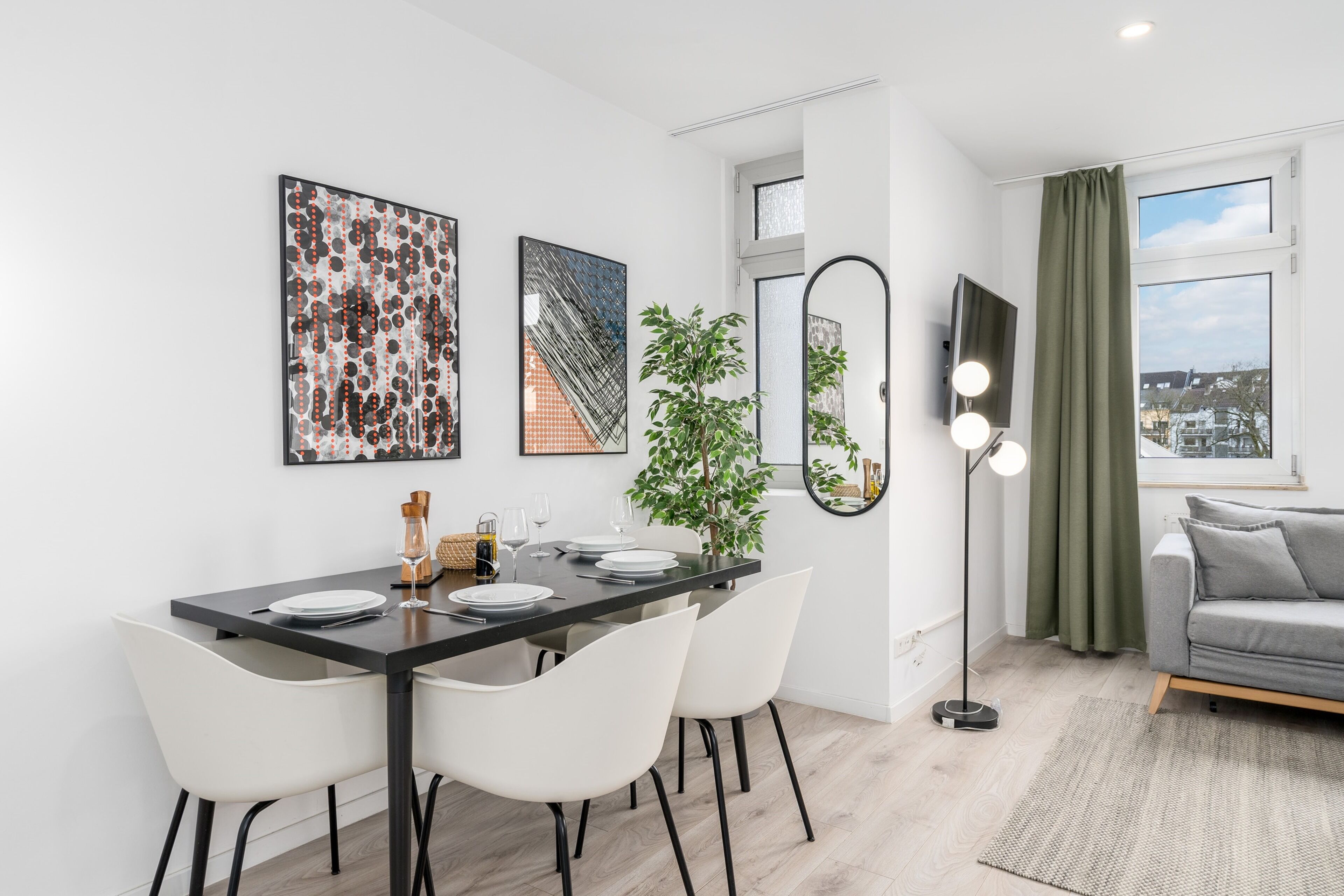 A modern dining area with a black table, white chairs, and a potted plant. There is a sofa and a window with curtains in the adjacent living area.