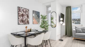 A modern dining area with a black table, white chairs, and a potted plant. There is a sofa and a window with curtains in the adjacent living area.