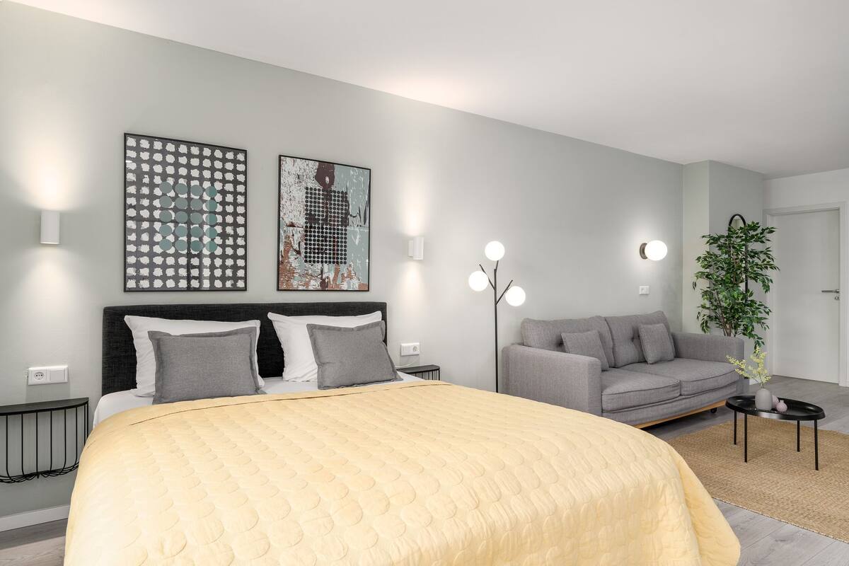 A modern bedroom with a large bed, a grey sofa, a small table, and two wall-mounted light fixtures.