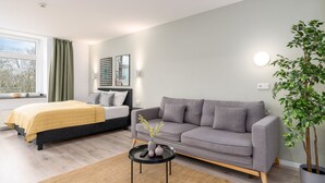 A modern bedroom with a bed, a grey sofa, a small table with a vase, and a potted plant. - BRIGHT Dortmund City Center (Dortmund)