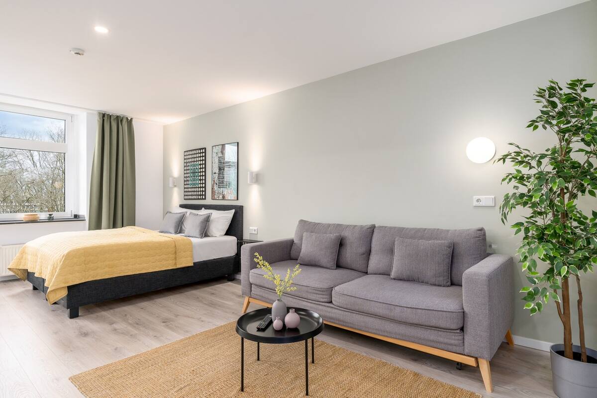 A modern bedroom with a bed, a grey sofa, a small table with a vase, and a potted plant.