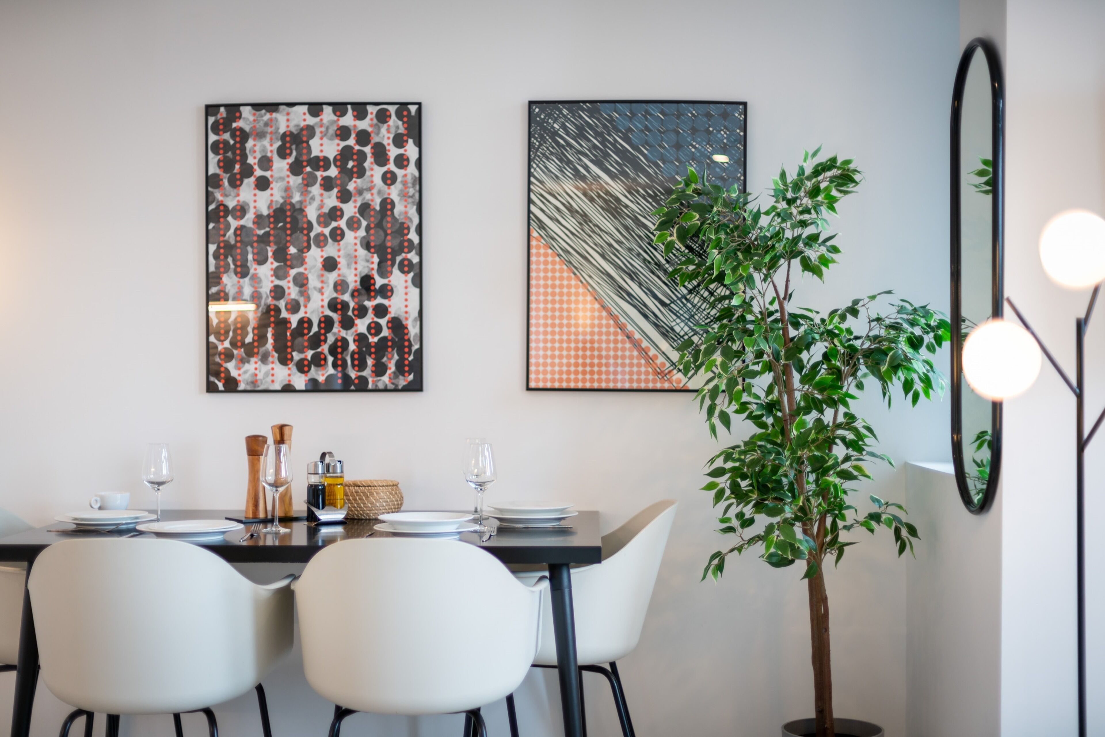 A dining area with a table set for two, two white chairs, a potted plant, and two framed artworks on the wall.