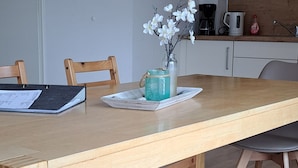 Une table à manger en bois, entourée de chaises, un vase avec des fleurs blanches, un carnet et une machine à café posés sur le comptoir.