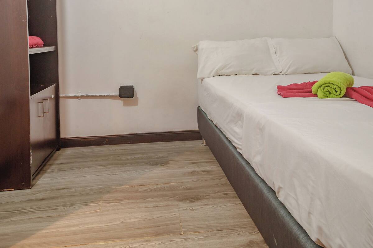Una habitación pequeña con una cama individual, suelo de madera, pared blanca, cama blanca y toalla verde, además de un ventilador de pie.