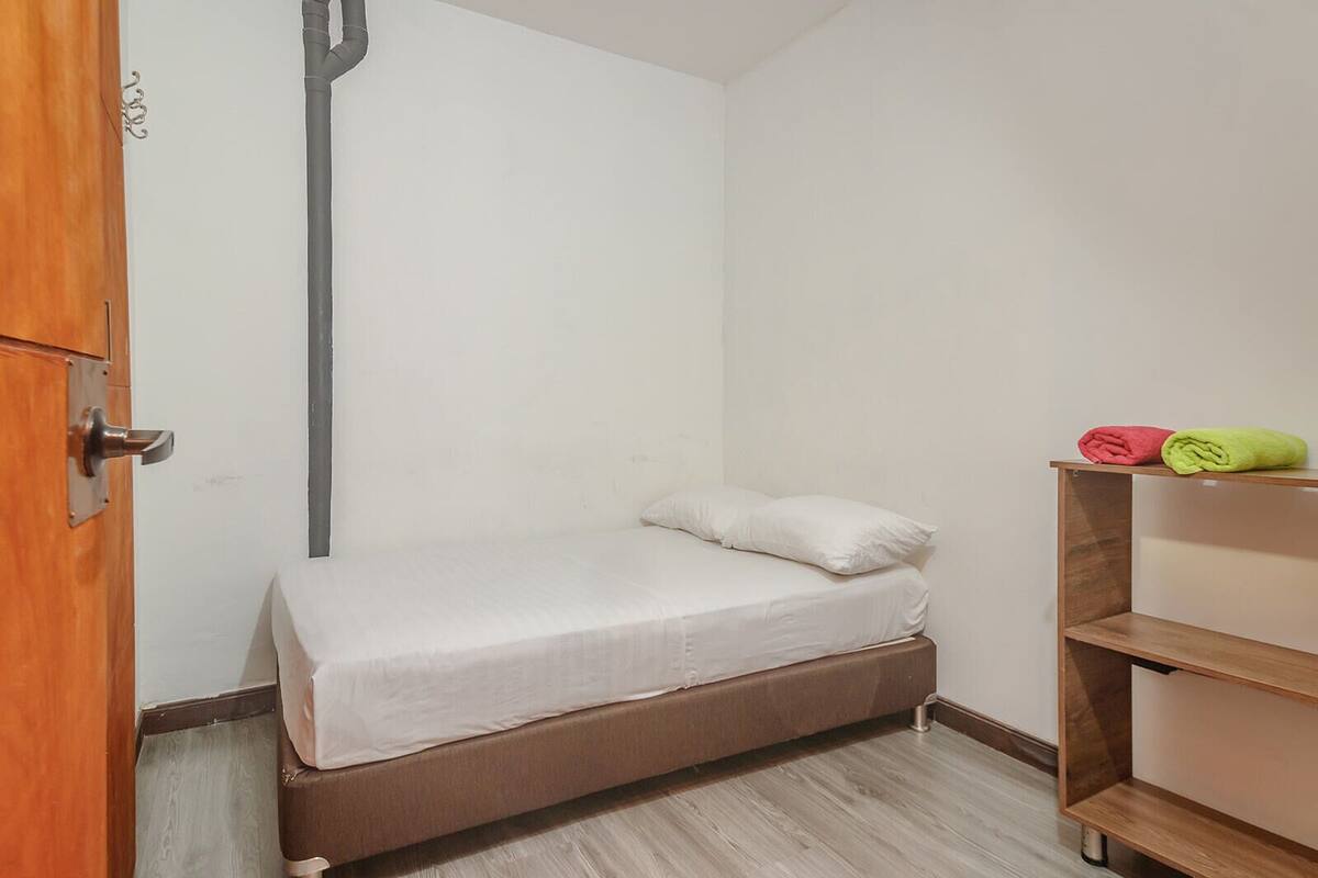 Una habitación pequeña con cama individual, puerta de madera, pared blanca y estante de madera con dos toallas enrolladas.