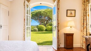 A bedroom with an open door leading to a garden, a bedside table with a lamp, and a framed picture on the wall.