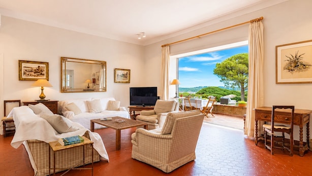 A spacious living room with a sofa, armchairs, a coffee table, and a television. There is a balcony with a table and chairs, and a large window offering a view of the outdoors.