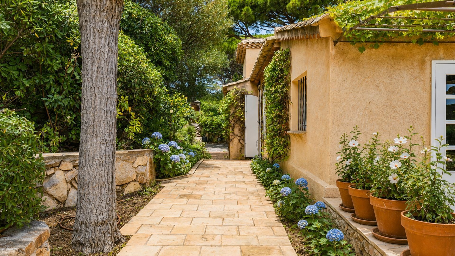 A stone pathway leading to a rustic building with a tiled roof, surrounded by lush greenery and flowering plants.