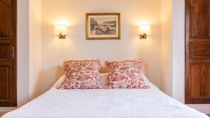 Una cama con dos almohadas estampadas, un cuadro de paisaje enmarcado encima y dos lámparas de pared a cada lado.