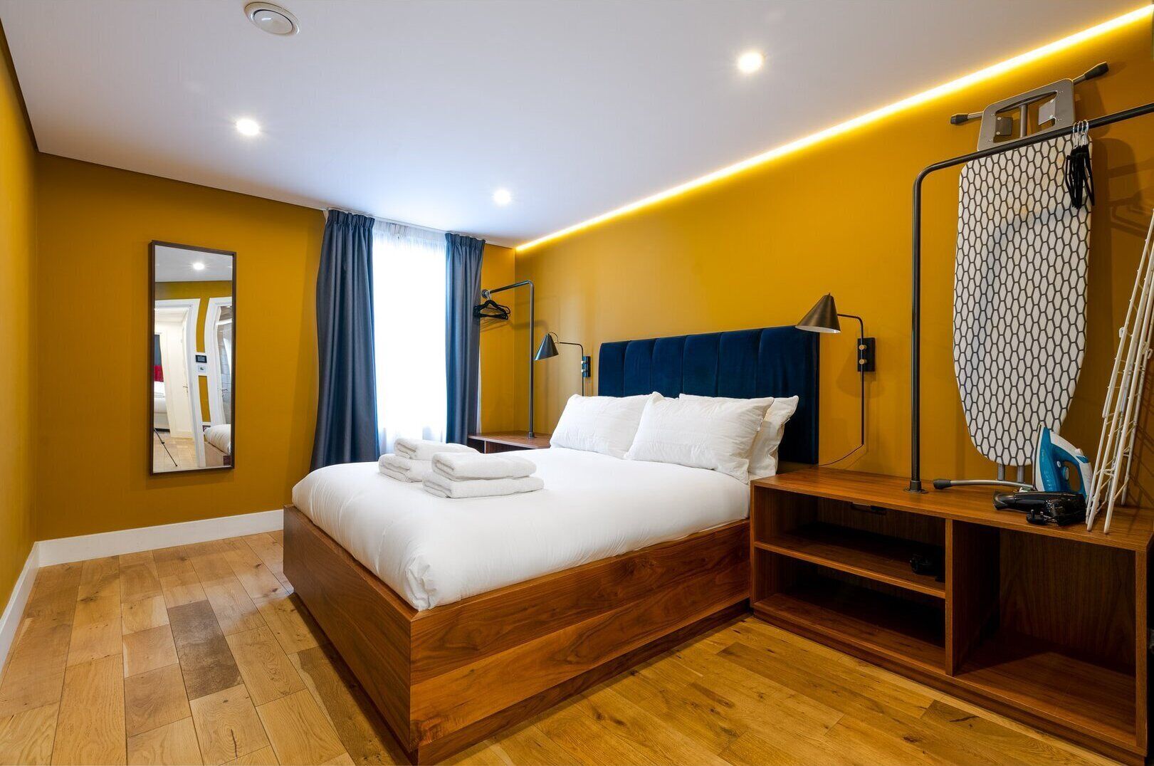 A modern hotel room with a wooden bed, a bedside table with an ironing board, a mirror, and a window with blue curtains.