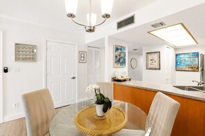 A modern kitchen with a glass dining table, chairs, a chandelier, and artwork on the walls. - Park Shore Resort 2nd Floor BLDG J #254 (Park Shore Resort)