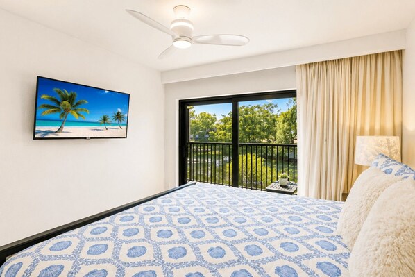Une chambre à coucher avec un lit, un ventilateur de plafond, une fenêtre donnant sur un paysage verdoyant et un tableau représentant une scène de plage.