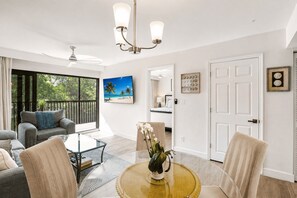 A modern living room with a glass coffee table, a sofa, and a dining area with chairs. There is a painting of a beach scene on the wall, and a door with a clock and decorative items.