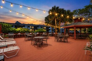 An outdoor patio area with multiple barbecue grills, tables, and chairs, surrounded by palm trees and string lights. - Park Shore Resort 2nd Floor BLDG J #254 (Park Shore Resort)