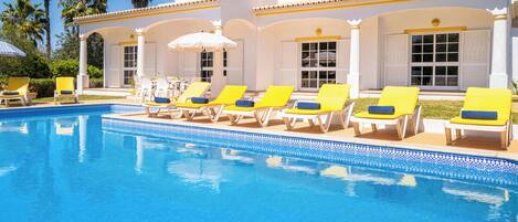 A pool area with sun loungers and umbrellas, surrounded by a building with white walls and columns.
