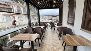 A restaurant with a stone-tiled floor, wooden tables and chairs, large windows with a view of a bridge and buildings, and a modern ceiling design. - Bujtina Apsus (Berat)