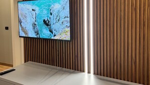 A modern interior with a flat-screen TV mounted on a wooden wall, a white console table, and a pendant light. - Bujtina Apsus (Berat)