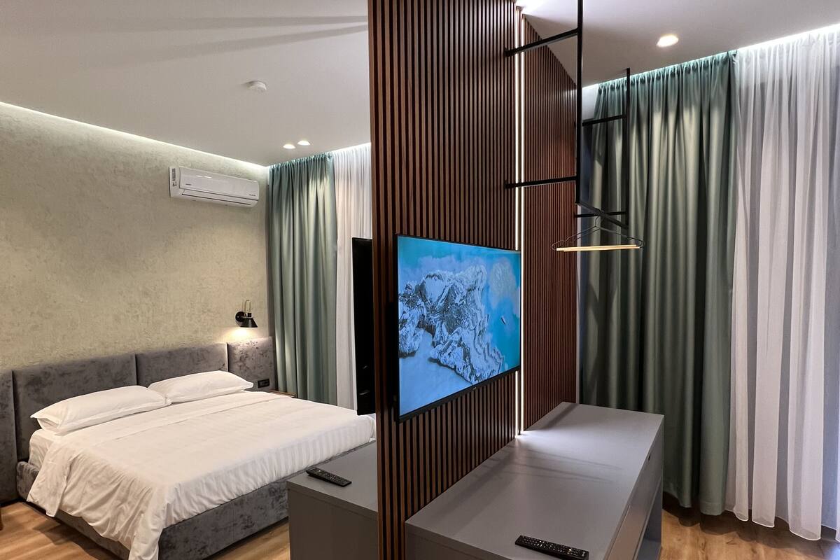 A modern hotel room with a large bed, a TV mounted on a wooden panel, a small desk, and a seating area with a sofa.