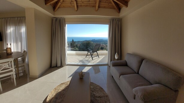 Een woonkamer met een bank, een salontafel en een eethoek met een tafel en stoelen. Er is een balkon met uitzicht op zee en een stoel.