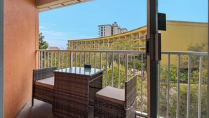 Un balcón con mobiliario de mimbre y una mesa de vidrio, con vistas a un edificio amarillo y vegetación.