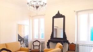 Un salon avec un canapé jaune, une table basse en bois et un lustre. Un grand miroir orne le mur, et une fenêtre est équipée de stores.