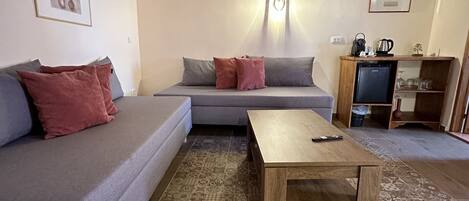 A cozy living room with a grey sofa, a wooden coffee table, patterned floor tiles, a wooden ceiling, and a small TV stand with a kettle.
