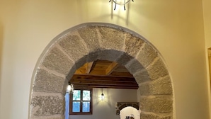 A cozy room with a stone archway leading to a bed and a small table, a window with a view, and a hanging light fixture. - Midgard Suites - Medieval Town (Rhodes)