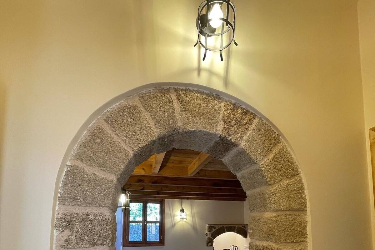 A cozy room with a stone archway leading to a bed and a small table, a window with a view, and a hanging light fixture.