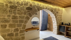 A bedroom with a stone wall, wooden ceiling, a bed with a blue and white striped bedspread, a wooden shelf, a small table with a kettle, and a mirror on the wall. - Midgard Suites - Medieval Town (Rhodes)