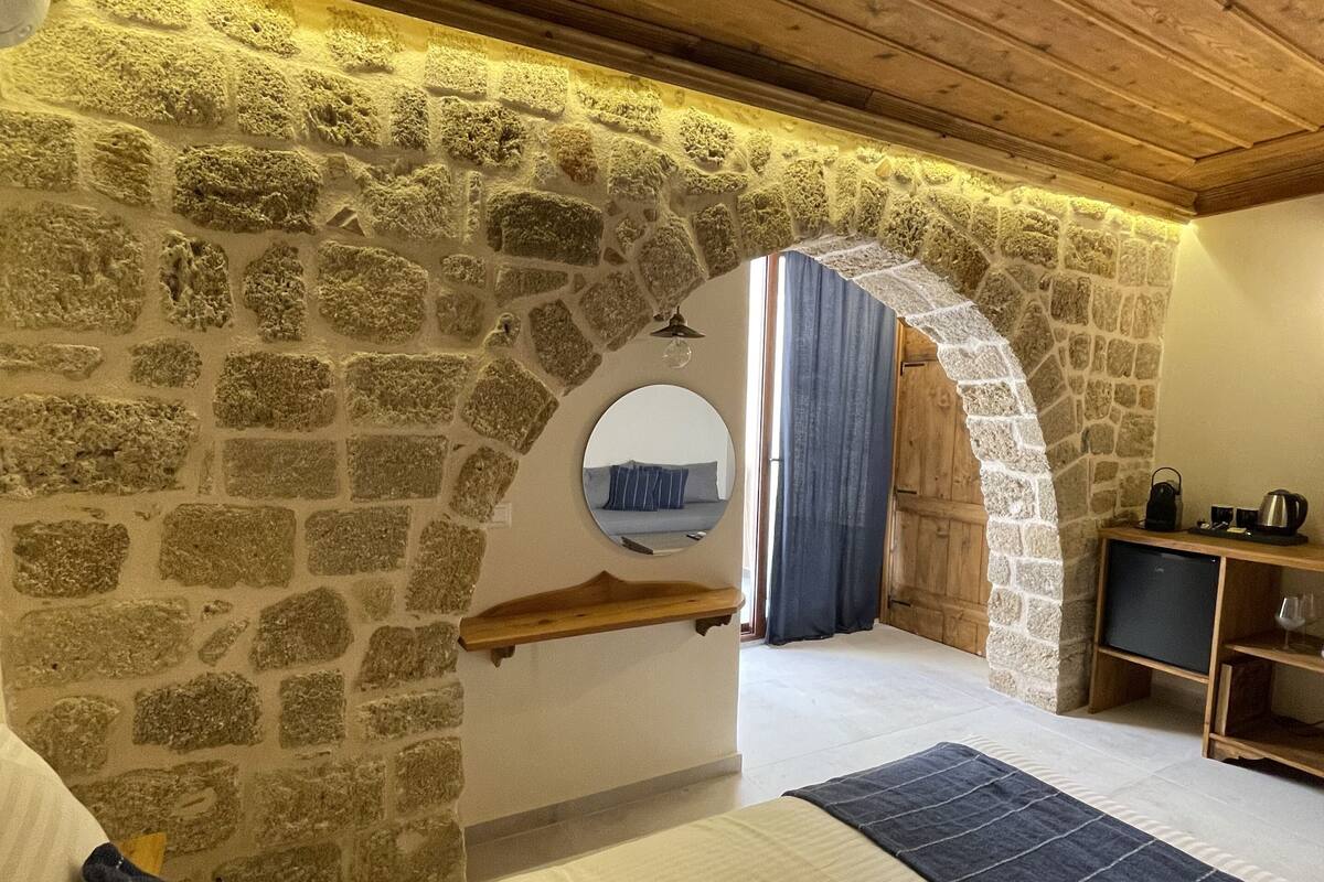 A bedroom with a stone wall, wooden ceiling, a bed with a blue and white striped bedspread, a wooden shelf, a small table with a kettle, and a mirror on the wall.