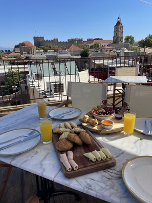 A terrace with a table set for breakfast, including croissants, pastries, and fruit, overlooking a historic cityscape with a prominent tower. - Midgard Suites - Medieval Town (Rhodes)