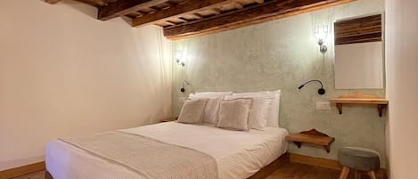 Une chambre avec un plafond en bois, un lit avec une literie blanche, une table de chevet, une lampe fixée au mur et un petit tabouret.