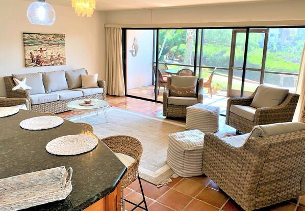 A spacious living area with a sofa, chairs, a dining table, and a view through large glass doors.
