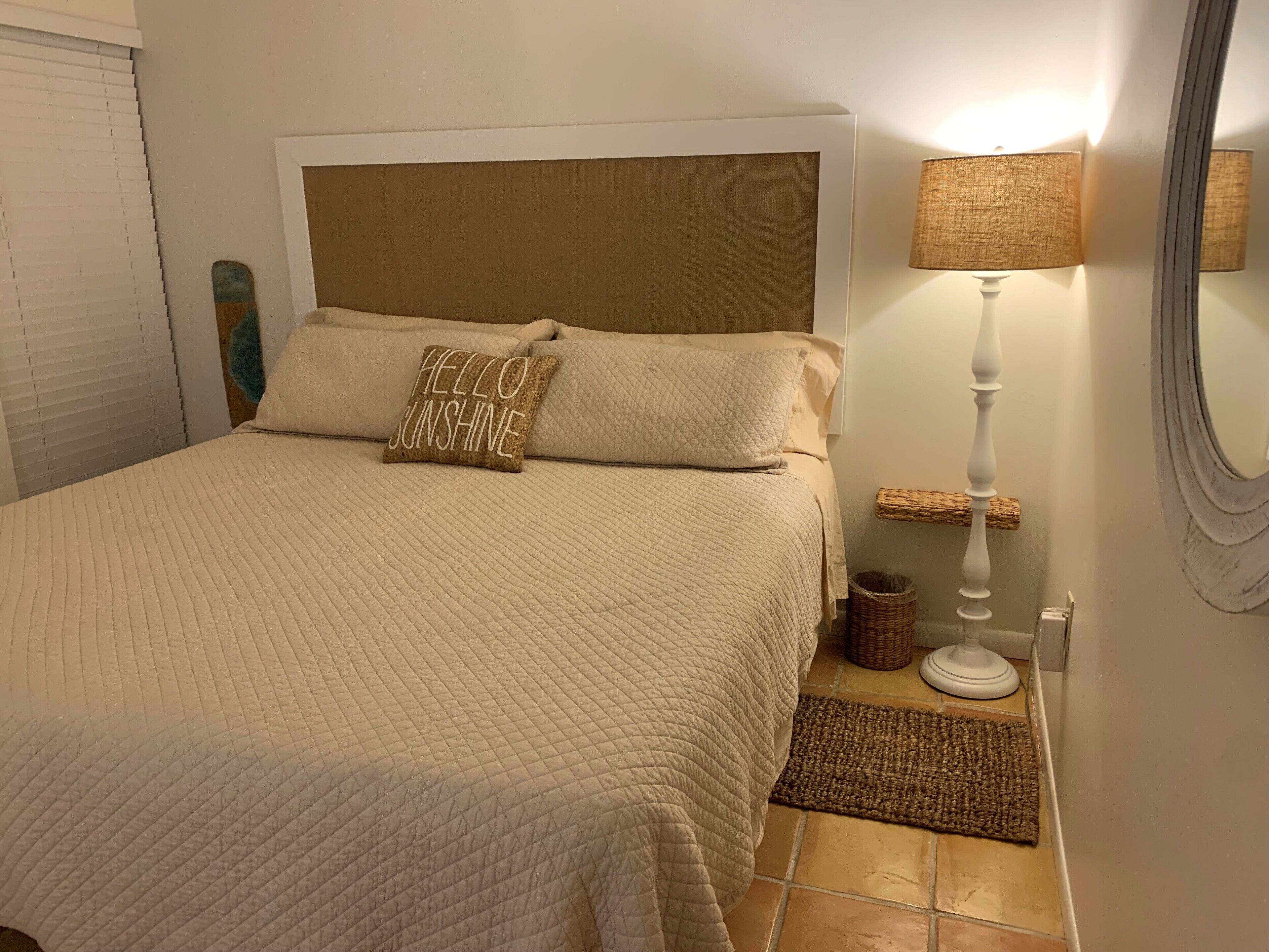 A neatly made bed with a quilted bedspread and two pillows, one with a text design. A bedside lamp and a small wicker basket are visible on the nightstand.