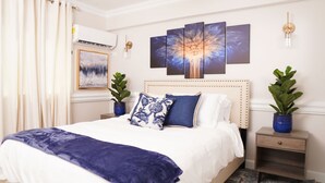A bedroom with a large bed, two nightstands, a wall-mounted air conditioner, a framed abstract painting, and a multi-panel artwork above the bed. - Modern Paradise Island Condo near Atlantis resort & beach (Nassau)