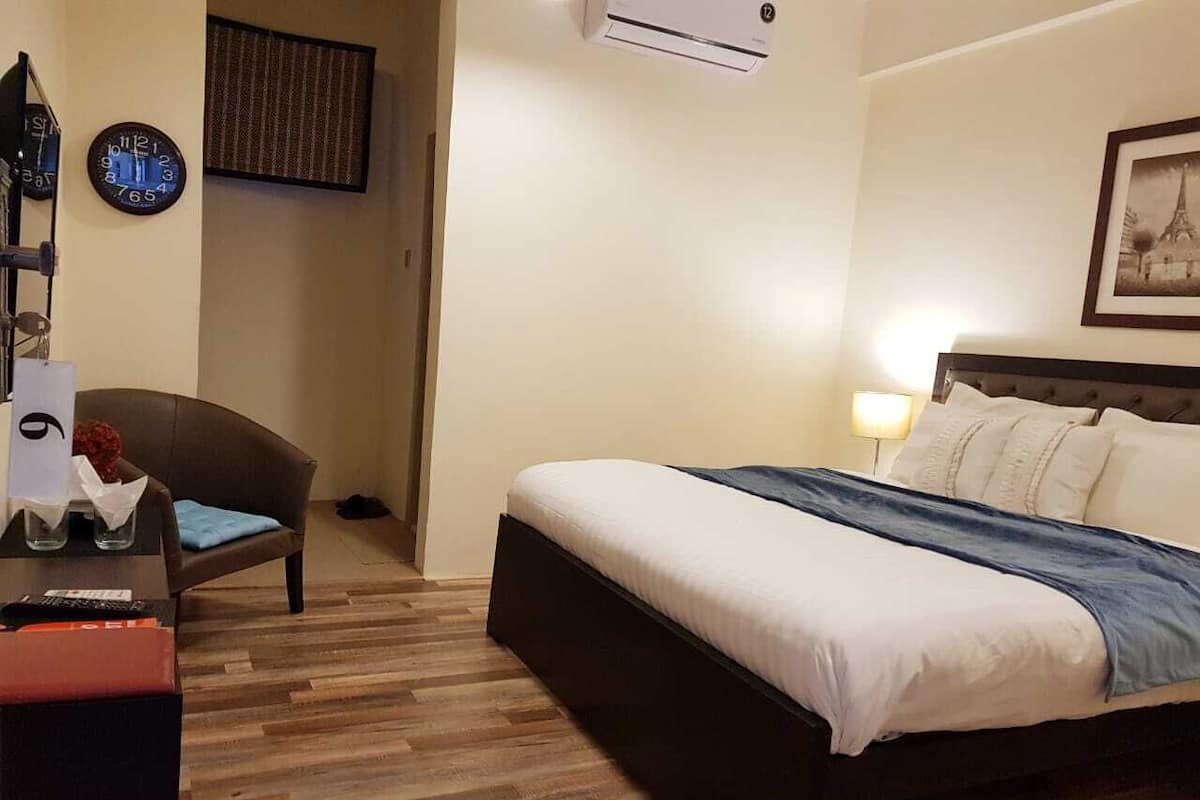 A hotel room with a bed, a desk, a chair, a clock, and an air conditioning unit.